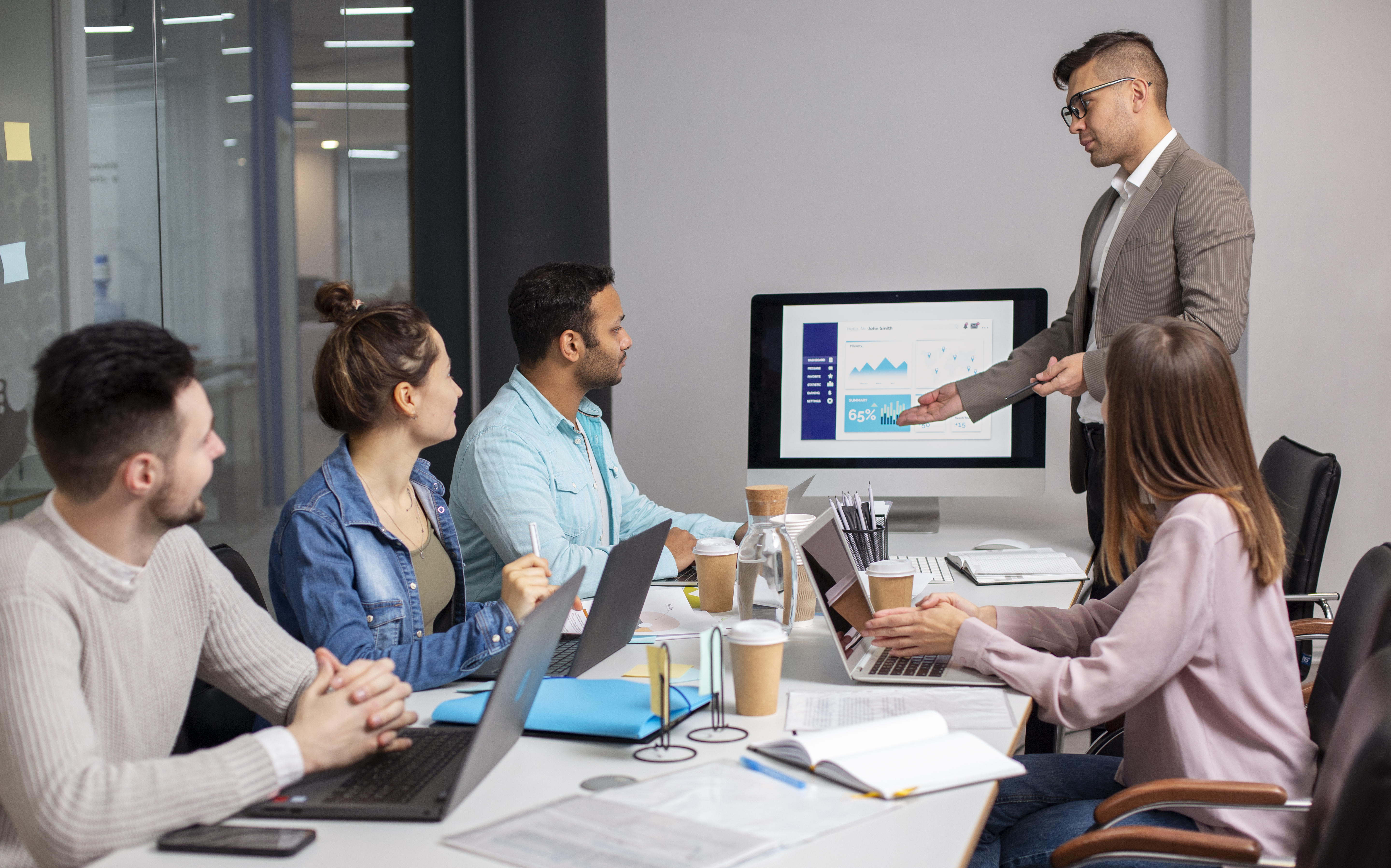 A group of office colleagues in a conference room sitting and listening to a man presenting data on screen