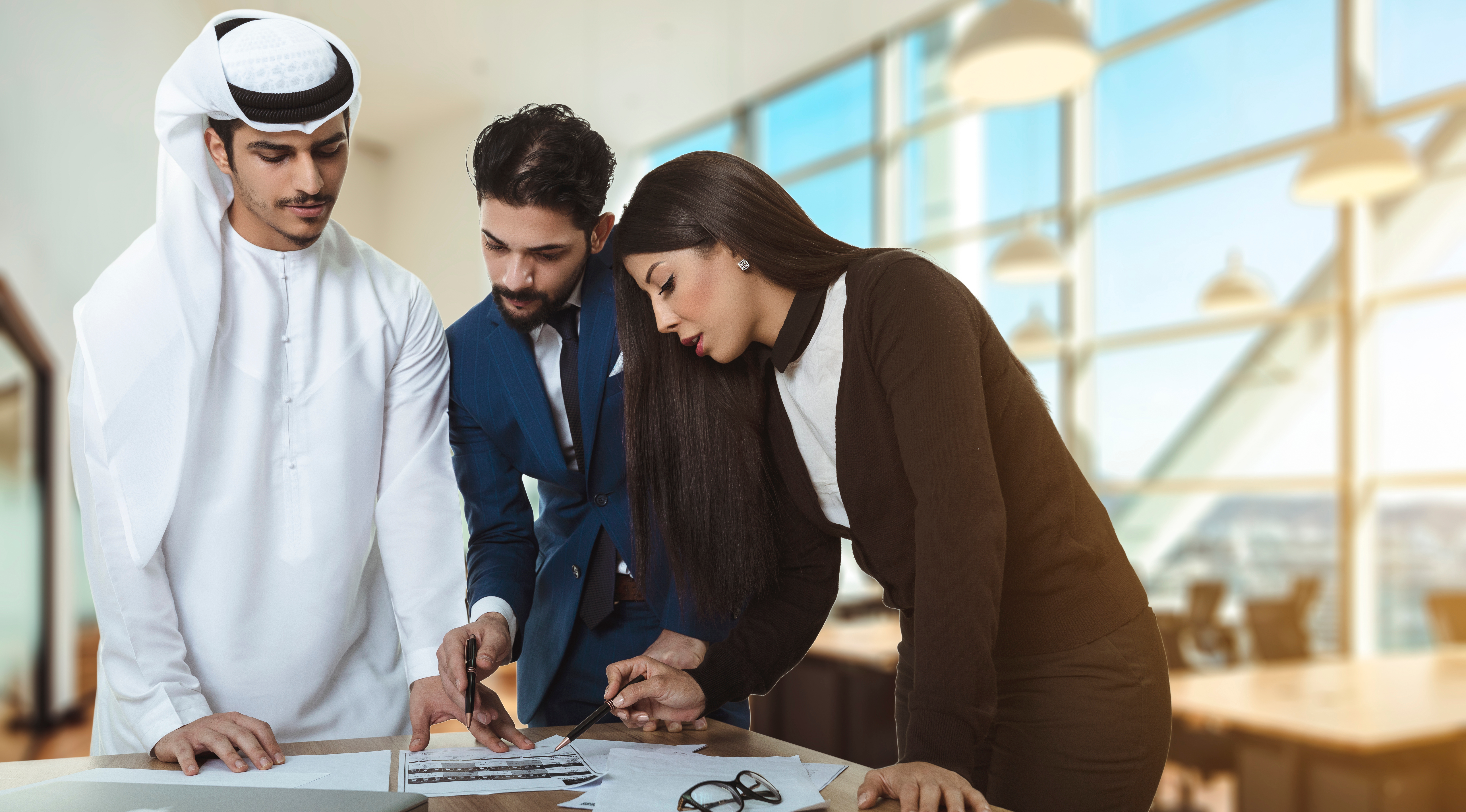 Arab man with a man in suit and a woman in black suit standing around a table discussing a paper report.