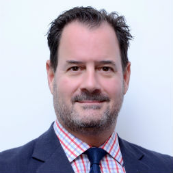 Smiling man in a suit and checkered shirt with a blue tie, posing against a plain background.