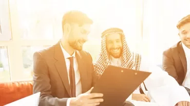 A diverse group of professionals smiling and discussing documents in a sunlit office.