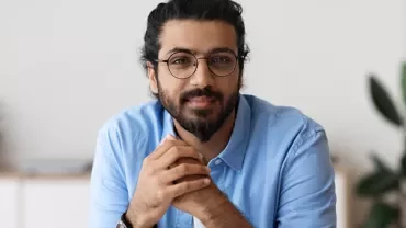 Man with glasses and beard in a blue shirt, sitting indoors.