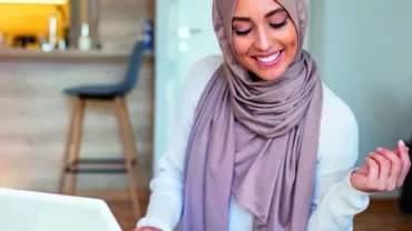 Woman in a hijab smiling at a laptop in a modern living space.