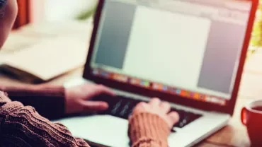 A person typing on a laptop with a blank document on the screen, a notepad, and a coffee cup on the wooden table.