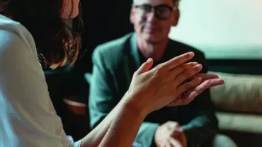 Close-up of hands gesturing during a conversation, with a person sitting on a sofa in the background.
