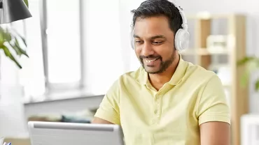 Man in yellow shirt wearing headphones, smiling while using a laptop.