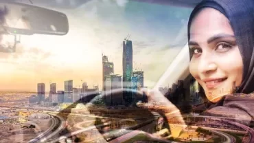 Woman in a headscarf driving with a city skyline in the background.