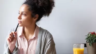 Woman in a sweater holding a pen and looking thoughtful, with juice and plant nearby.