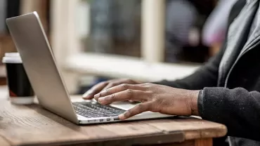 Person typing on a laptop in a cafe with a takeaway coffee cup nearby.