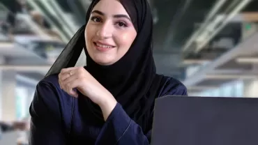 Smiling woman in hijab working on a laptop in an office.