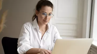 A woman in a white shirt and glasses working on a laptop at a desk.
