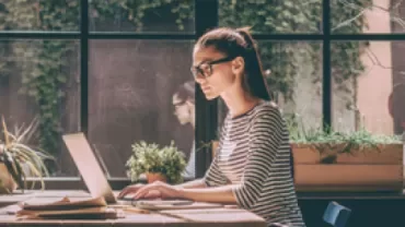 Woman in glasses working on a laptop near a window with plants.