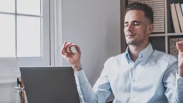 Man in blue shirt meditating at a desk with a laptop.