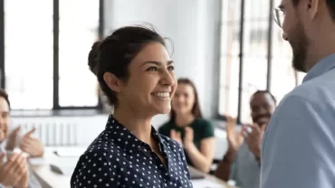 Woman smiling at a man in an office with colleagues clapping.
