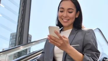 Woman in a suit smiling at her phone while going up an escalator outdoors.