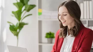Smiling woman in a red blazer working on a laptop in a bright office.