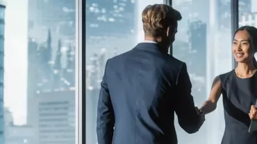 Two business professionals shaking hands in a glass-walled office with a cityscape view.