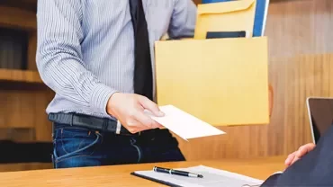 A man in office handing over a resignation letter with a box of his belongings in another hand to the manager sitting. 