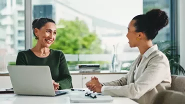 Two women engaged in discussion at a table with a laptop open between them