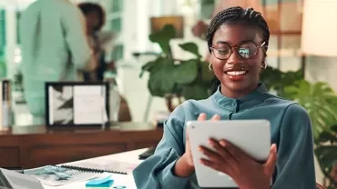 Woman standing in the office on her ipad with her laptop behind her
