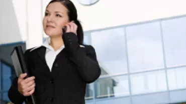 Businesswoman in black suit talking on a phone and holding a folder outside a modern office building.