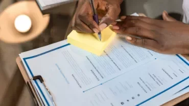 Person writing on sticky notes near a resume on a clipboard.
