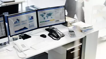 Office desk with dual monitors displaying charts and data, phone, and keyboard.