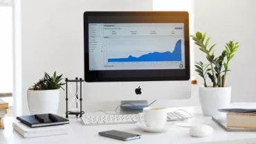 Desktop with a monitor displaying a rising graph, surrounded by plants and office supplies.