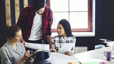 Three people in a meeting room discussing architectural plans around a table.