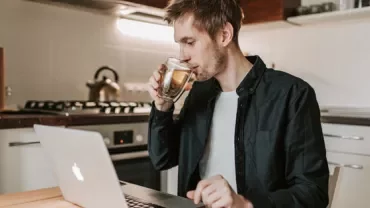 Man drinking coffee while working on a laptop in a kitchen.