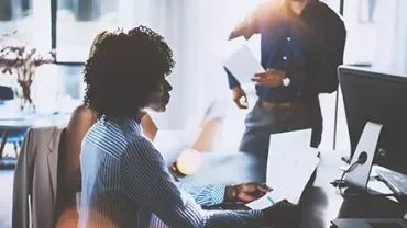 Team having a discussion in a bright office room, with a focus on a woman using a laptop.
