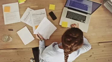 Woman at desk with laptop and papers, working and writing notes.