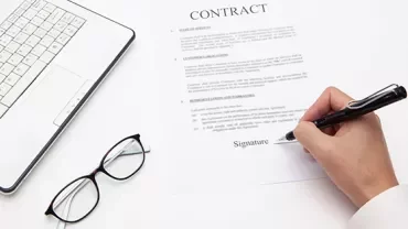 Person signing a contract beside a laptop and glasses.