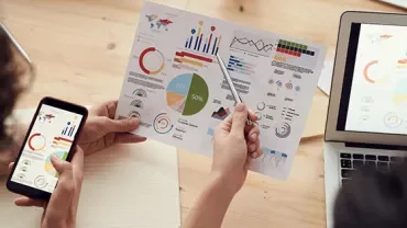 Person analyzing charts on paper with phone and laptop at desk.