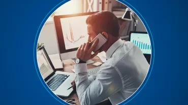 Man at desk analyzing graphs on computer screens, talking on phone.