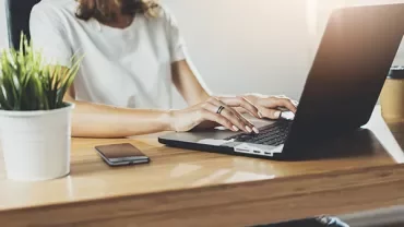 Person typing on a laptop at a wooden desk with a plant and smartphone nearby.