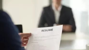 Person holding a resume during a job interview, facing a blurred figure in the background.