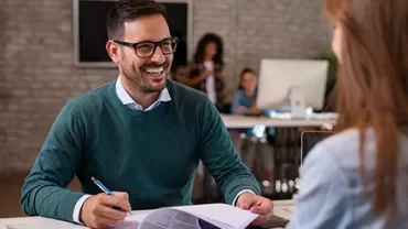 Smiling man in a green sweater holds papers while talking to a woman in an office setting.