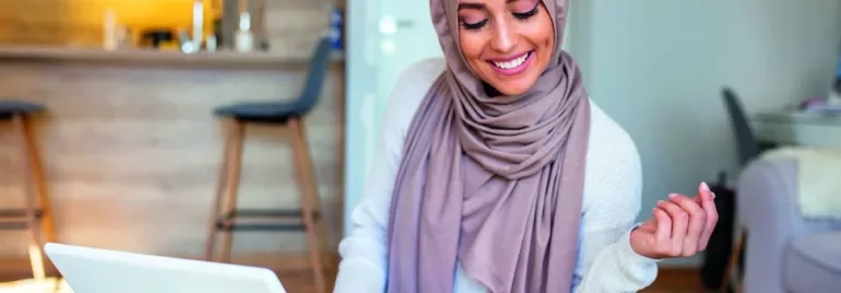 Woman in a hijab smiling at a laptop in a modern living space.