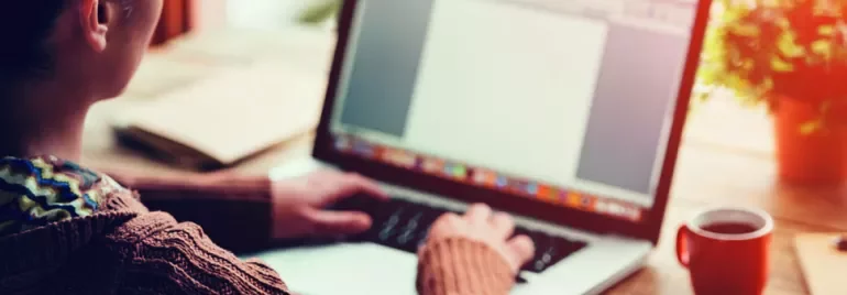 A person typing on a laptop with a blank document on the screen, a notepad, and a coffee cup on the wooden table.