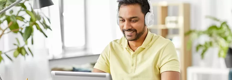 Man in yellow shirt wearing headphones, smiling while using a laptop.