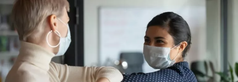 Two women wearing masks greet each other with an elbow bump.