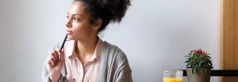 Woman in a sweater holding a pen and looking thoughtful, with juice and plant nearby.