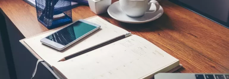 A wooden desk with a planner, smartphone, pencil, coffee cup, and a small plant.