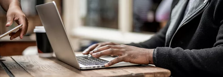 Person typing on a laptop in a cafe with a takeaway coffee cup nearby.