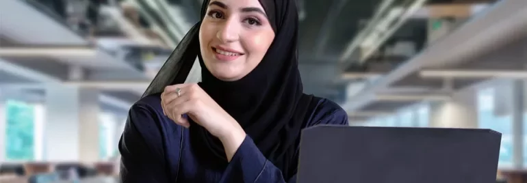 Smiling woman in hijab working on a laptop in an office.