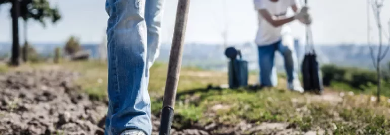 Person in jeans digs soil with a shovel, another person in background planting saplings.