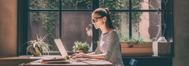 Woman in glasses working on a laptop near a window with plants.