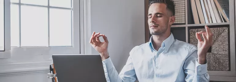Man in blue shirt meditating at a desk with a laptop.