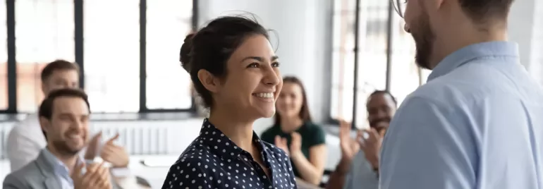 Woman smiling at a man in an office with colleagues clapping.