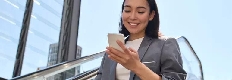Woman in a suit smiling at her phone while going up an escalator outdoors.